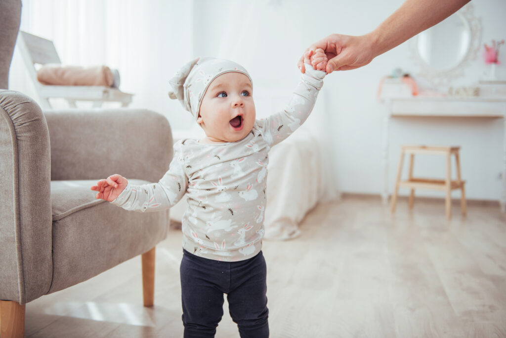 Ein Baby bei den ersten Gehversuchen an der Hand eines Elternteils im Wohnzimmer